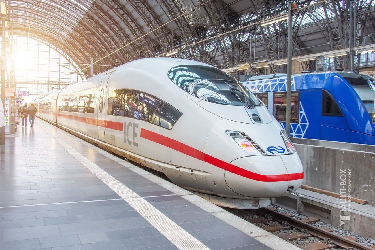 Ein eleganter weiß-grauer Hochgeschwindigkeitszug in einem modernen Bahnhof mit gewölbtem Glasdach. Der Zug ist ein „ICE“ und hat rote Akzente. Auf dem angrenzenden Gleis ist ein zweiter Zug in Blau zu sehen. Sonnenlicht fällt durch den Bahnhof.
